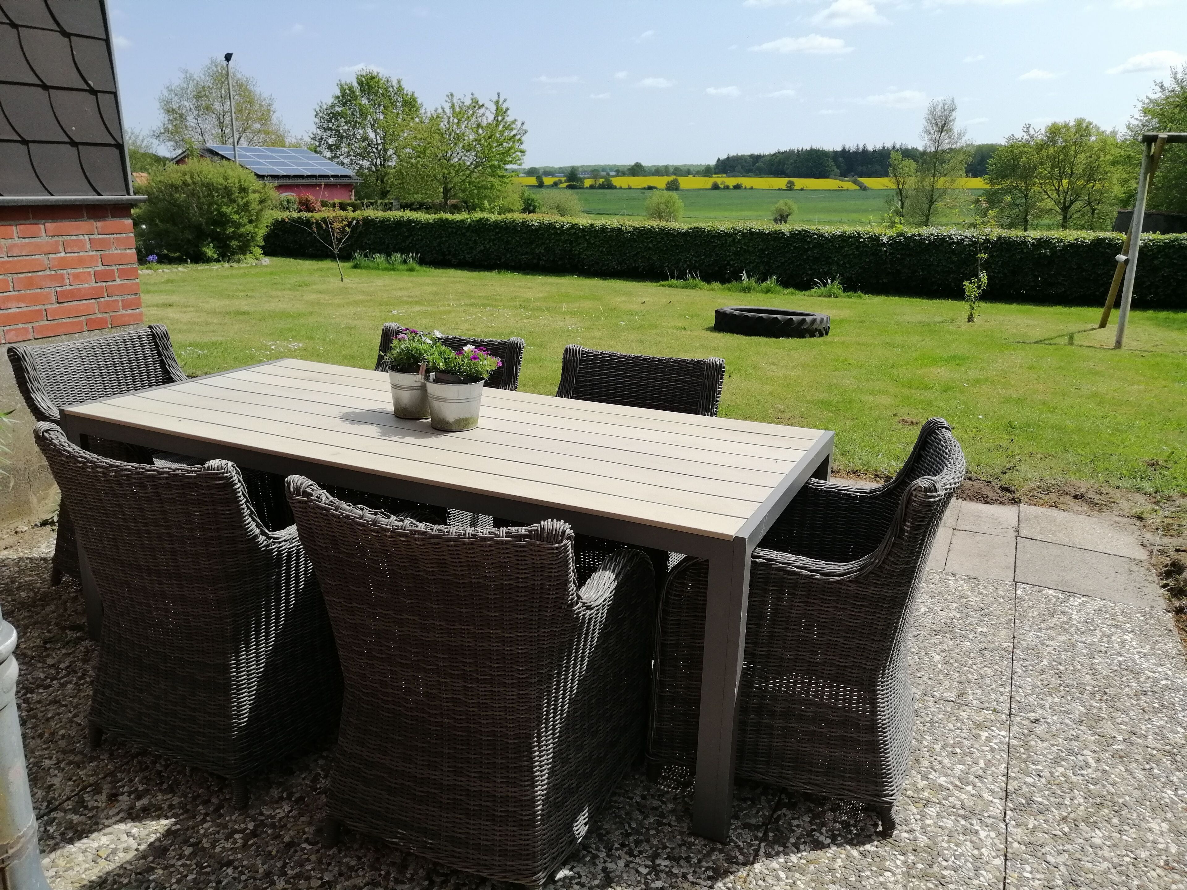 Um pátio externo com mesa e cadeiras de vime, rodeado por um jardim e vista para campos.