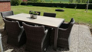 Une terrasse extérieure avec une table et des chaises en osier, entourée d’un jardin et offrant une vue sur les champs.