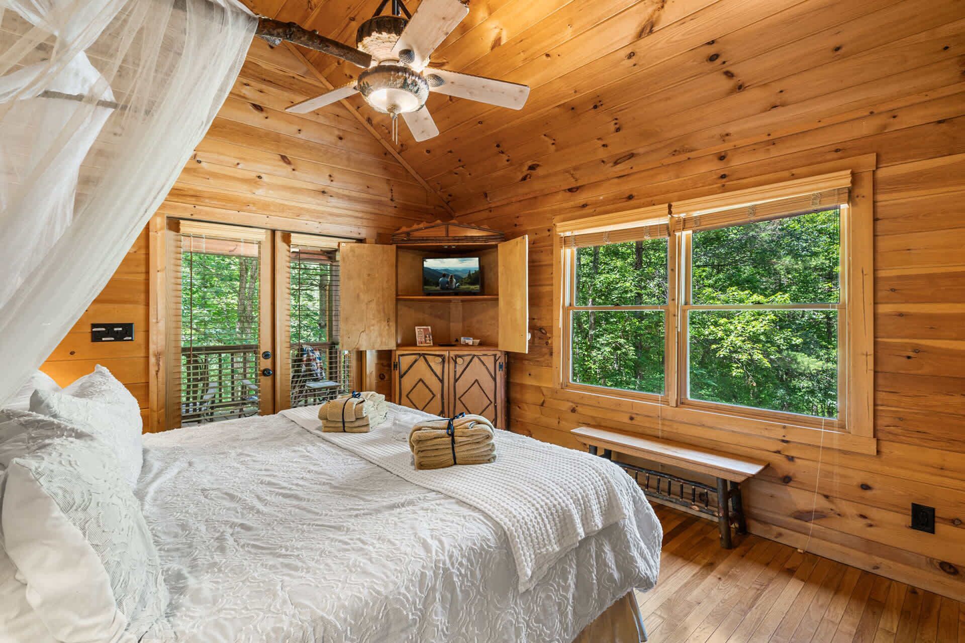 A wooden cabin bedroom with a bed, a ceiling fan, a television, and large windows.