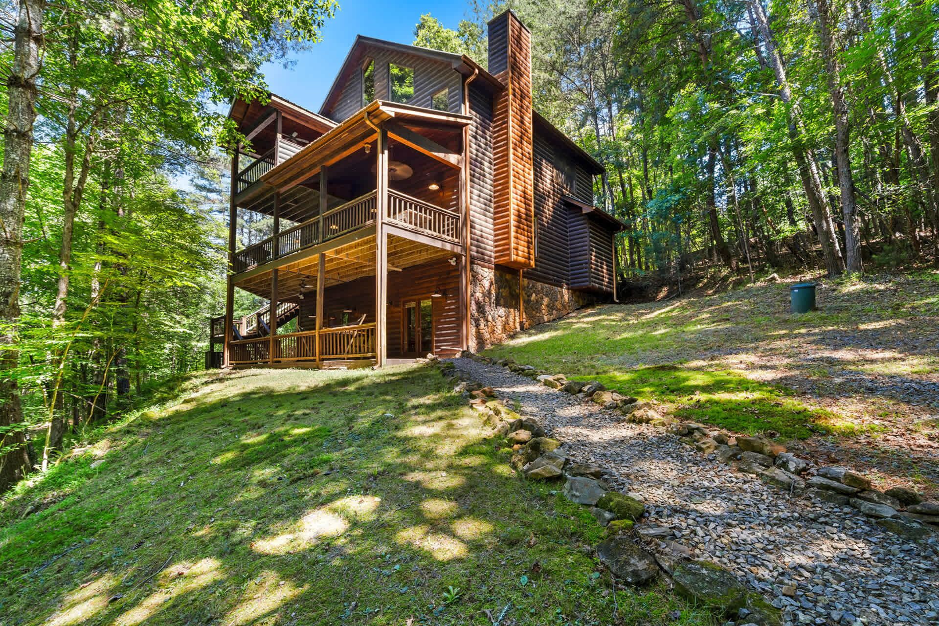 A two-story log cabin with a balcony and a stone chimney, surrounded by a forest.