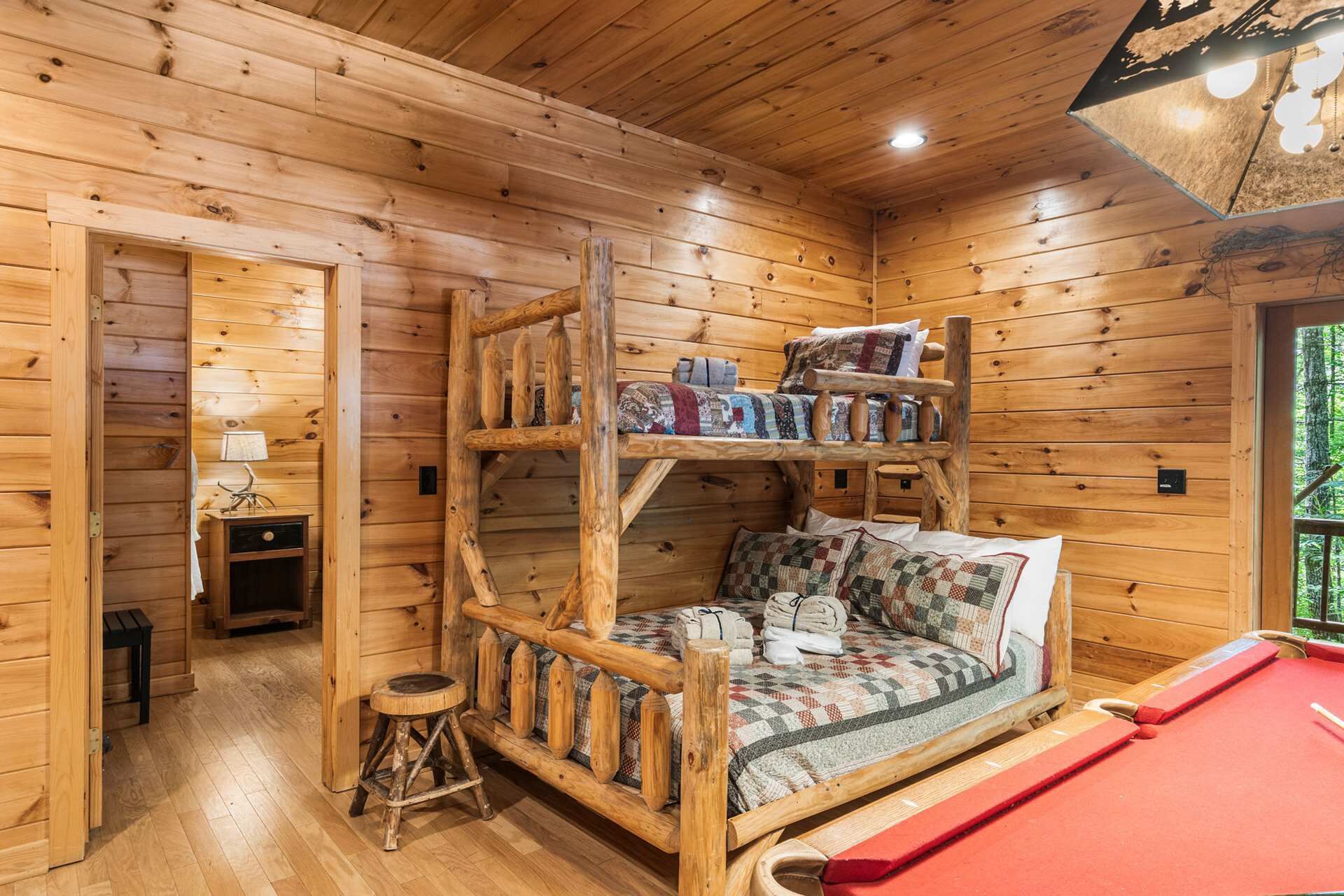 A wooden cabin interior with a bunk bed, a small table with a lamp, and a red pool table.