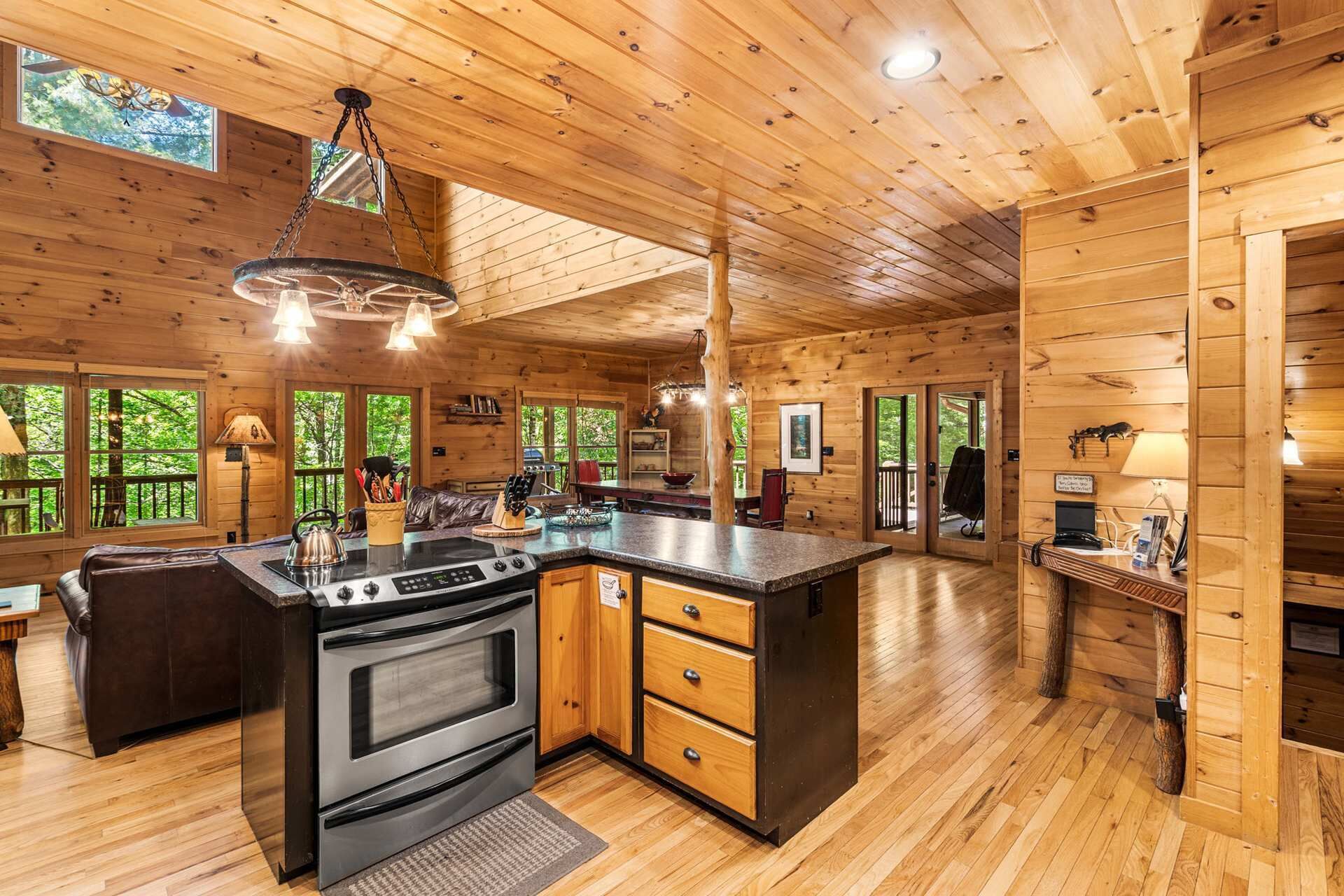 A wooden cabin interior with a kitchen, dining area, and a large window overlooking greenery.