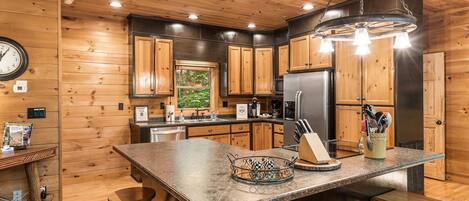 A kitchen with wooden cabinets, a stainless steel refrigerator, a large island with stools, and a hanging light fixture.