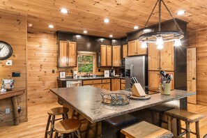 A kitchen with wooden cabinets, a stainless steel refrigerator, a large island with stools, and a hanging light fixture.