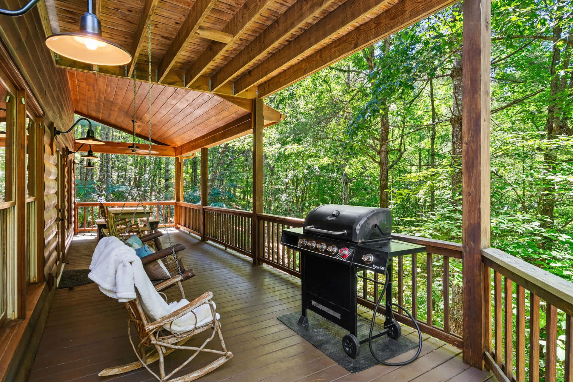A covered porch with a grill, wooden deck, rocking chair, and a view of the forest.
