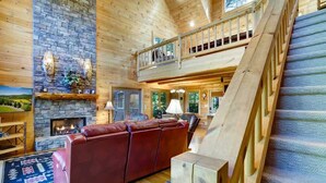 A spacious living area with a fireplace, a red leather sofa, and a wooden staircase. - Hot Tub & Scenic Mountain Views, Game Room (Mineral Bluff)
