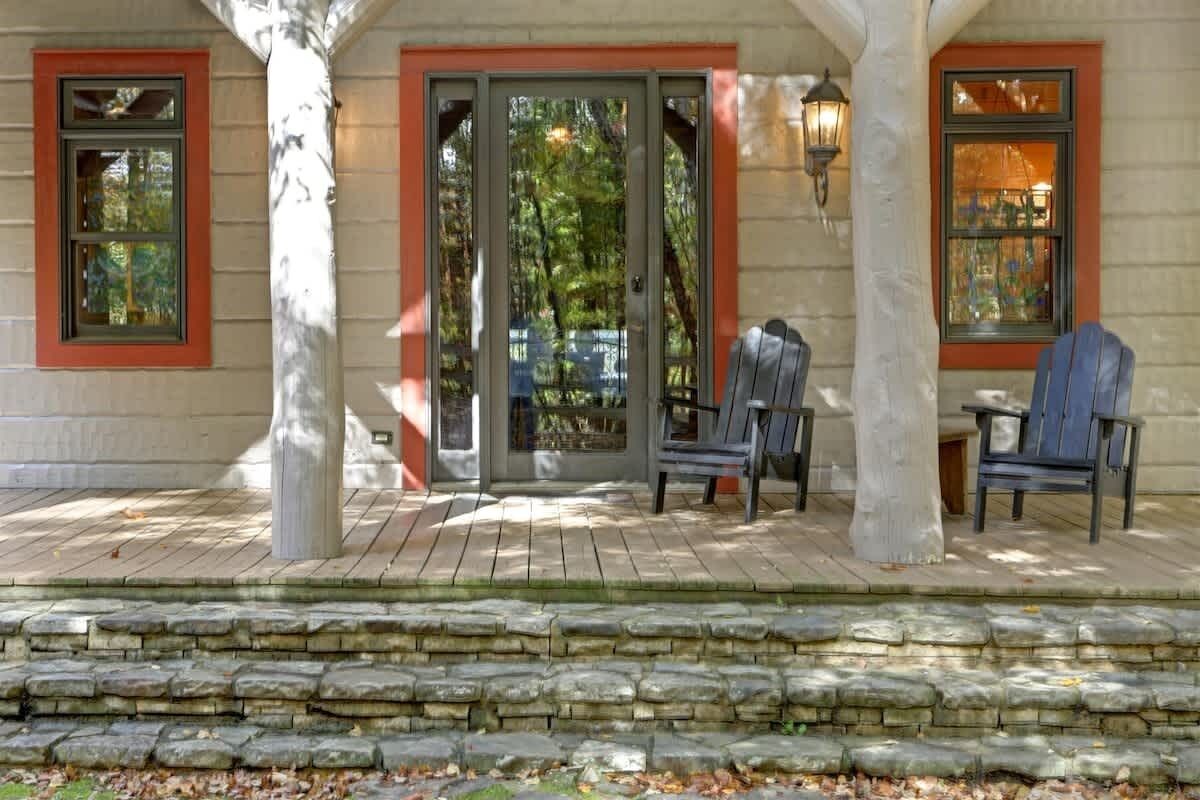 Un porche cubierto con escalones de piedra, dos sillas Adirondack y una terraza de madera.