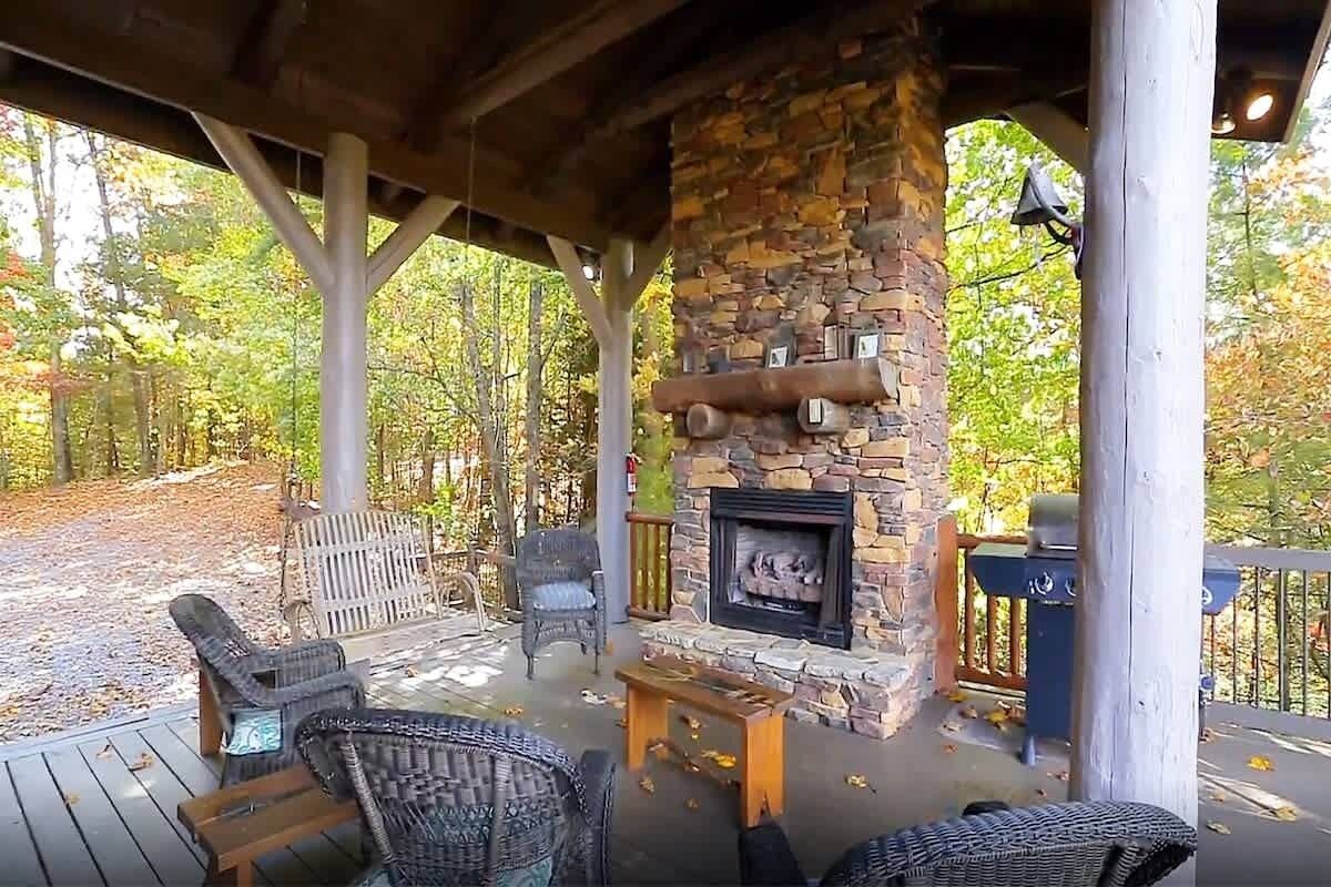 A covered outdoor seating area with wicker chairs, a stone fireplace, and a view of the forest.