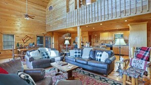 A spacious cabin interior with a living area featuring a large blue sofa, a wooden coffee table, and a fireplace. A kitchen is visible in the background, and a balcony overlooks the living space. - Stunning Mountain Sunset Views, screened-in HotTub (Mineral Bluff)