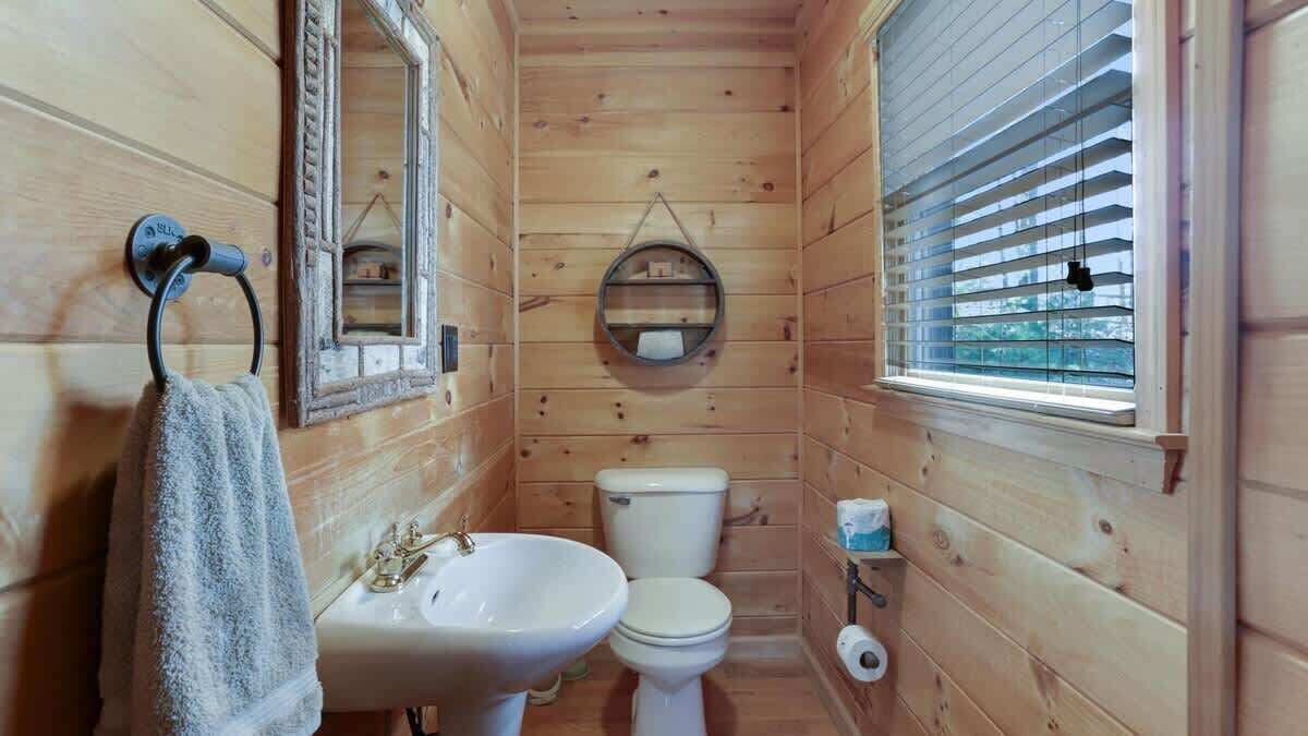 A wooden-paneled bathroom with a sink, toilet, and window with blinds.
