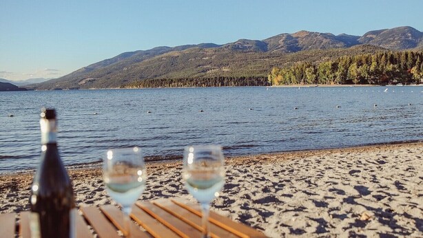 Une table avec une bouteille et des verres, placée sur une plage de sable, avec un lac et des montagnes en arrière-plan.