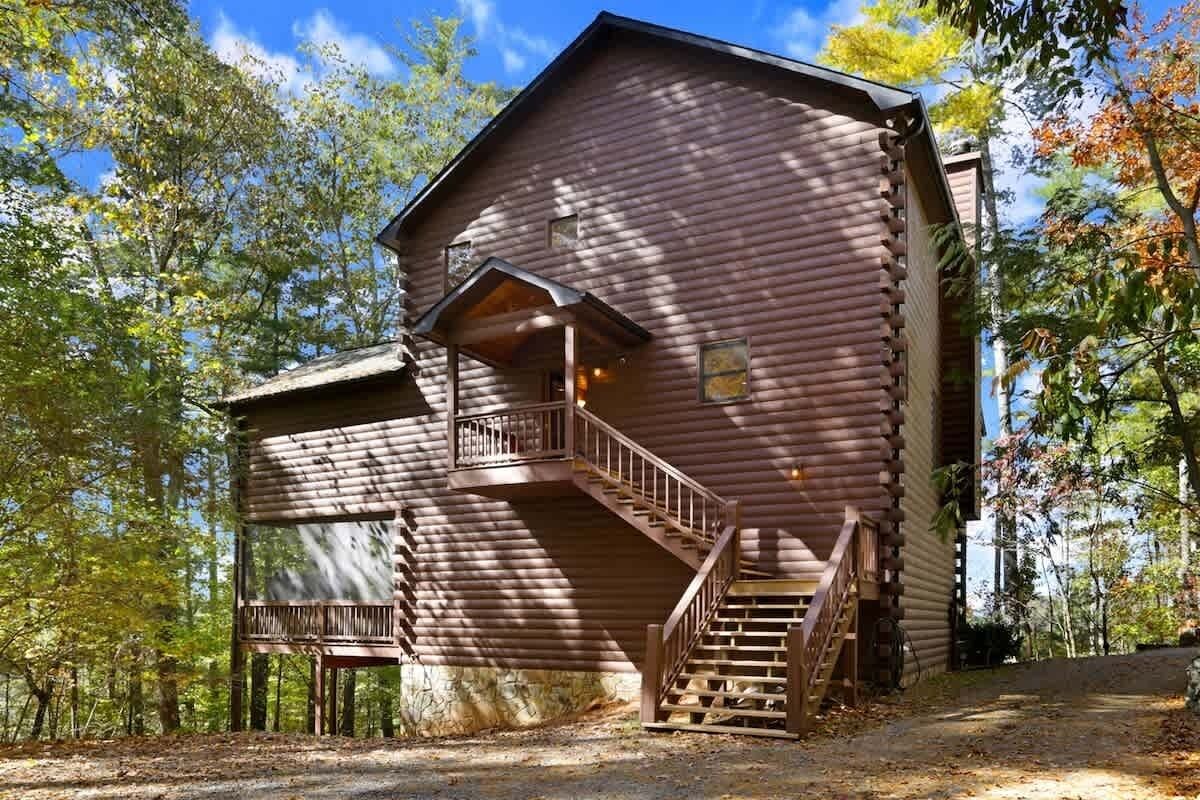 A rustic log cabin with a wooden staircase leading to a balcony, surrounded by trees.