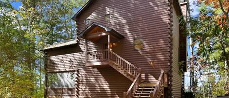 A rustic log cabin with a wooden staircase leading to a balcony, surrounded by trees.