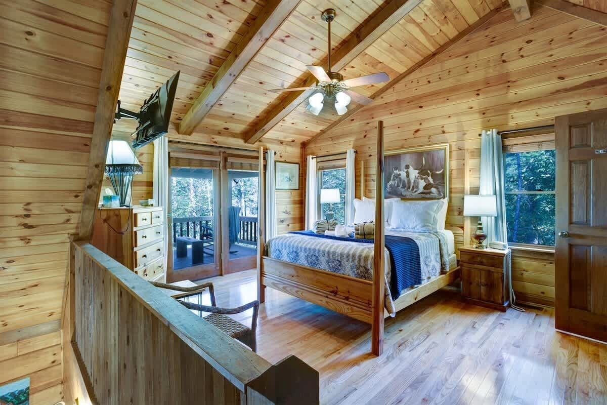 Un dormitorio de cabaña de madera con una cama con dosel, un ventilador de techo y ventanas grandes.