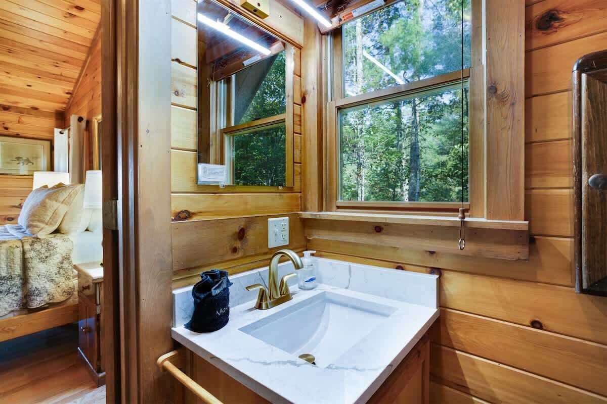 Innenansicht einer Holzhütte mit einem weißen Waschbecken, einem Spiegel und einem Fenster mit Blick auf den Wald.