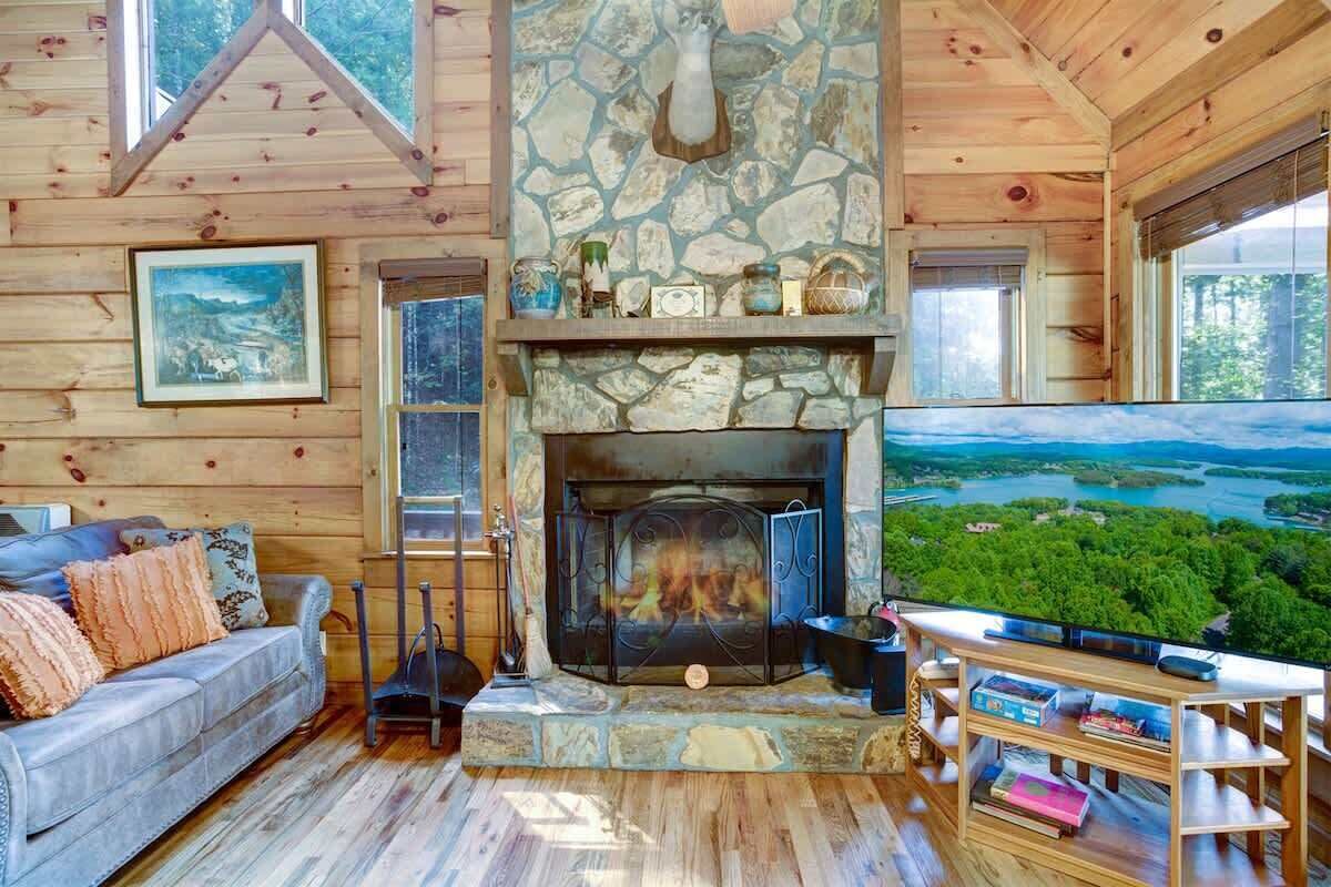 Un intérieur de chalet chaleureux avec une cheminée en pierre, une grande fenêtre donnant sur une forêt et un coin salon confortable.