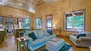 Ein großzügiger Wohnbereich mit einer großen Couch, einem Couchtisch und einem Esstisch. Es gibt eine Küchenzeile mit einer Mikrowelle und einem Kühlschrank. Das Zimmer verfügt über große Fenster mit Blick ins Freie.