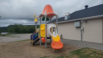 Un parque infantil con un tobogán y estructuras para escalar, frente a un edificio de techo gris y revestimiento color beige.