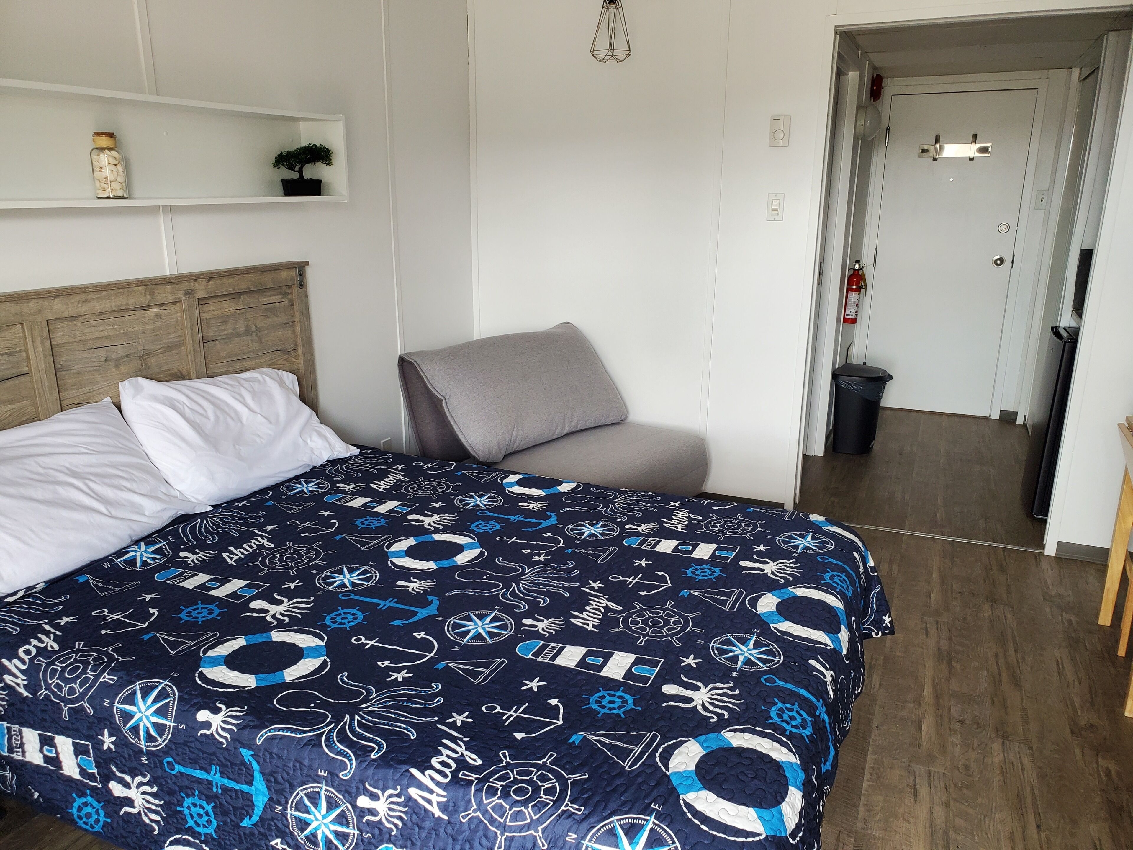 Een slaapkamer met een bed, een grijze stoel, een houten hoofdeinde, een plank met een plant en een pot, en een deur naar een gang.