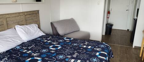 Un dormitorio con cama, una silla gris, cabecera de madera, estante con una planta y un frasco, y una puerta que da a un pasillo.