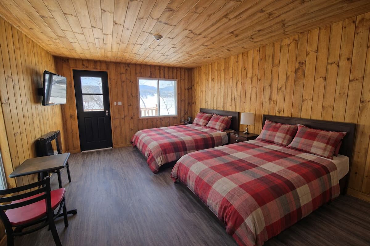 Ein Zimmer mit Holzwänden und -decke, zwei Betten mit Karo-Bettwäsche, einem Schreibtisch mit Stuhl, einem Fernseher und einem Fenster mit Bergblick.
