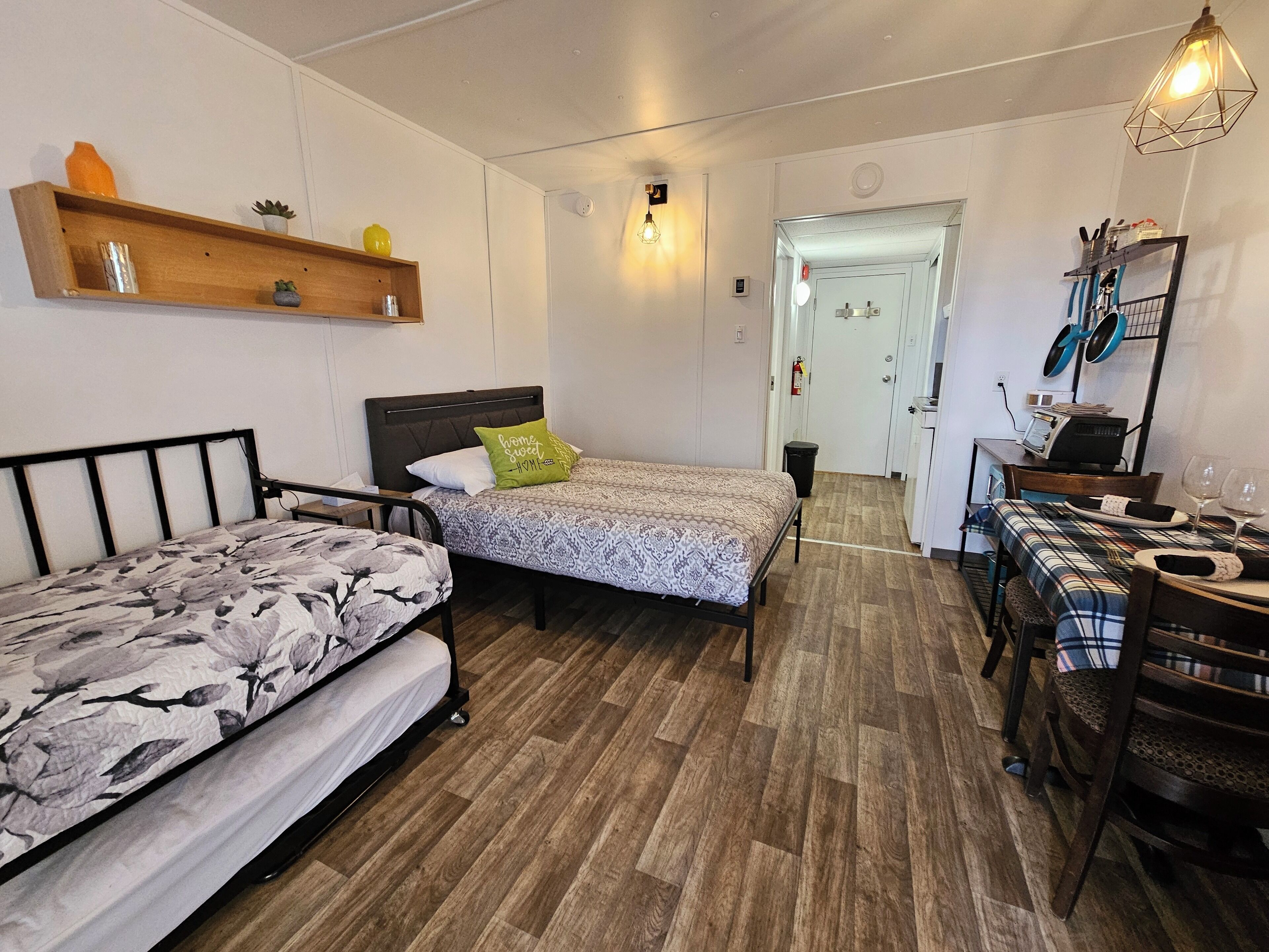 A compact bedroom with two beds, a wooden shelf, a small table, and a dining area with chairs.