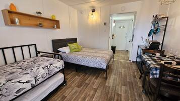 A compact bedroom with two beds, a wooden shelf, a small table, and a dining area with chairs.