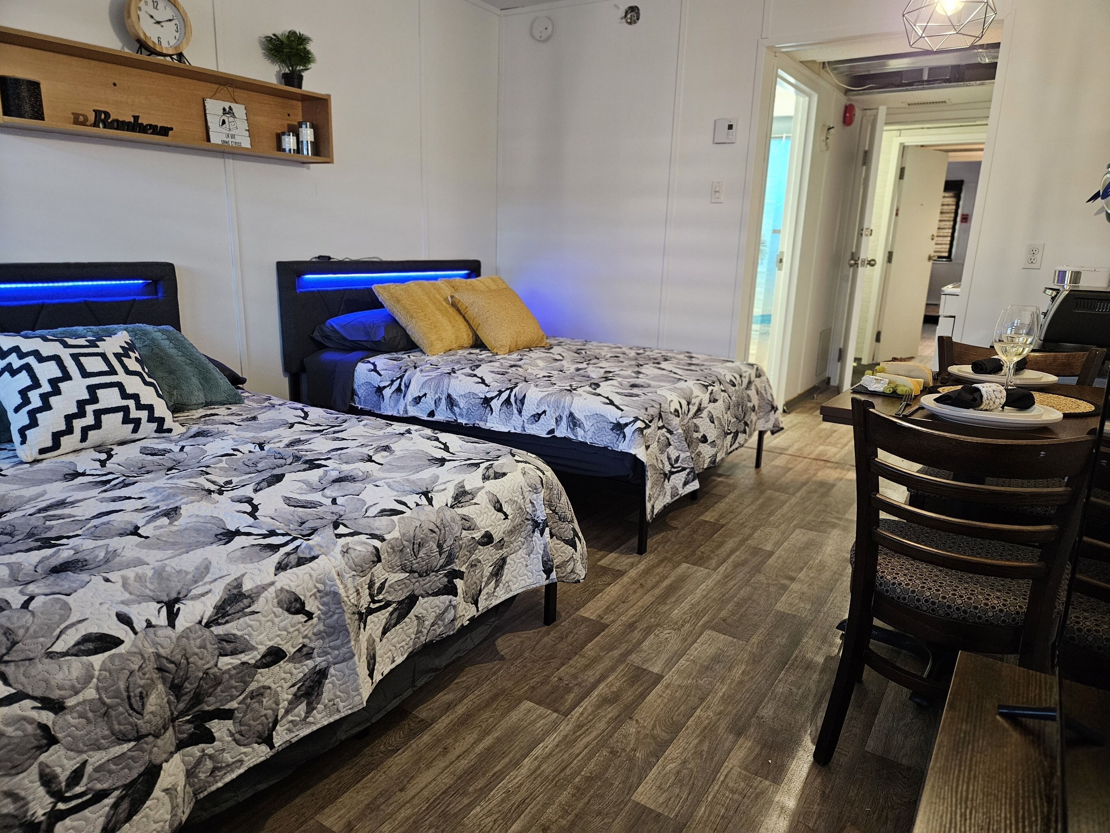 Hotellihuone, jossa on kaksi sänkyä, puinen pääty, pieni hylly, jossa on tavaroita, sekä ruokailutila, jossa on pöytä ja tuolit.