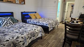 Habitación de hotel con dos camas, un cabecero de madera, un estante con objetos y una zona de comedor con mesa y sillas.