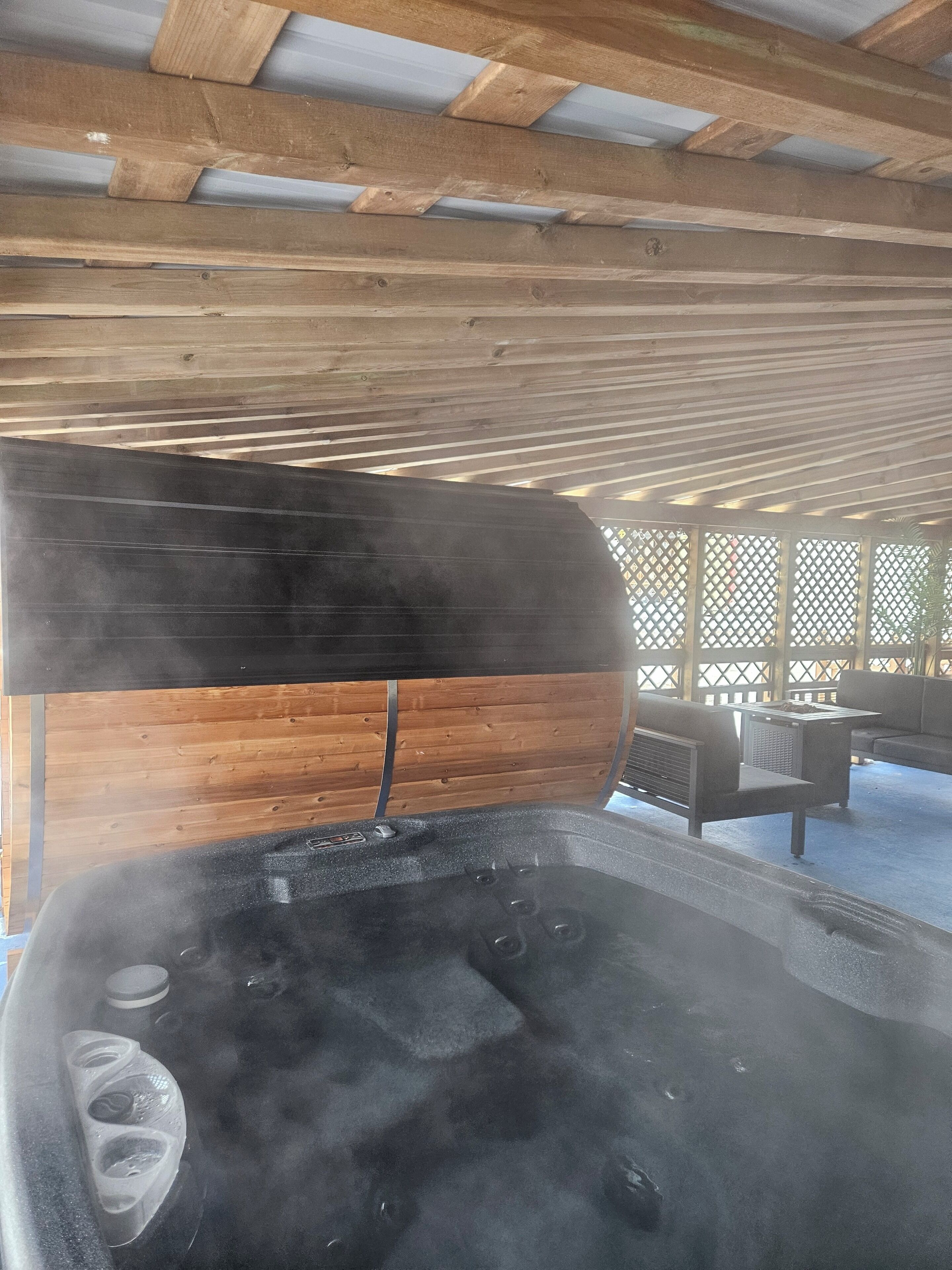 A hot tub with steam rising, wooden beams overhead, and a wooden fence in the background.