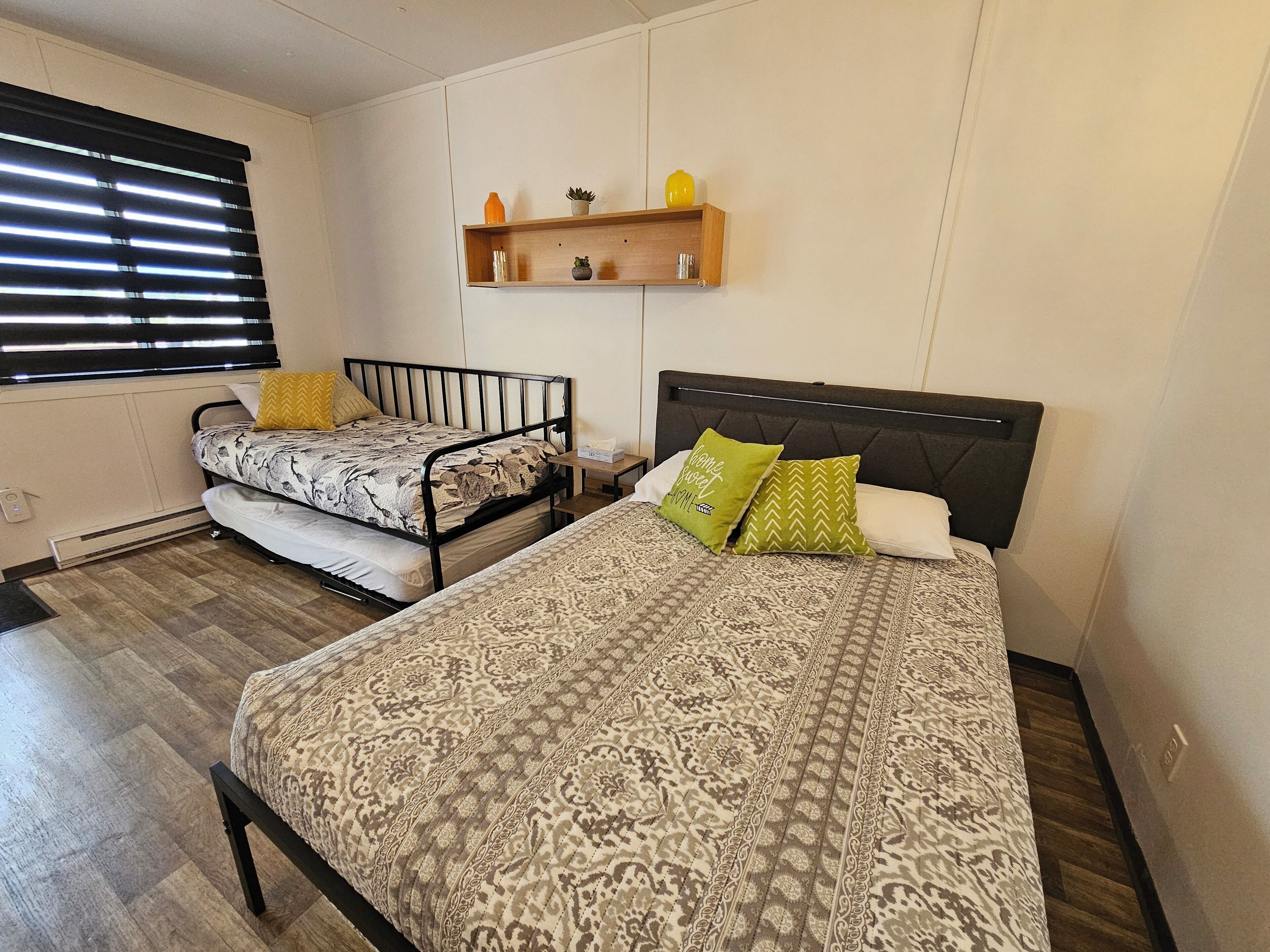 A bedroom with two beds, a wooden floor, a wall-mounted shelf with decorative items, and a window with blinds.