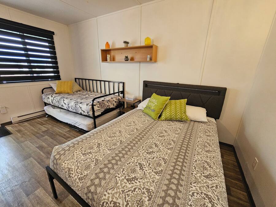 Une chambre avec deux lits, un plancher en bois, une étagère murale décorée de divers objets, et une fenêtre avec des stores.