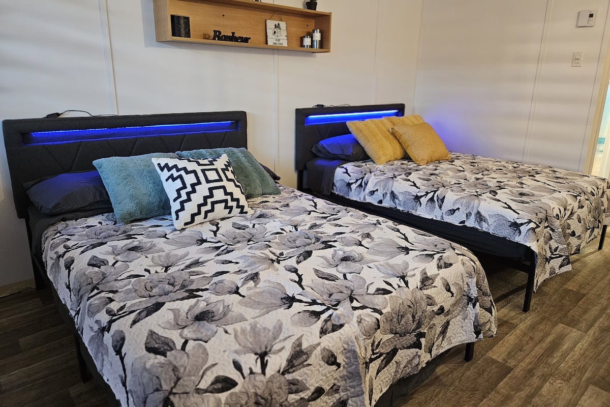 Ein Schlafzimmer mit zwei Betten, jeweils mit floralem Bettbezug und einem Kopfteil mit blauer LED-Beleuchtung. Darüber befindet sich ein Regal mit einer Uhr und einer kleinen Pflanze.