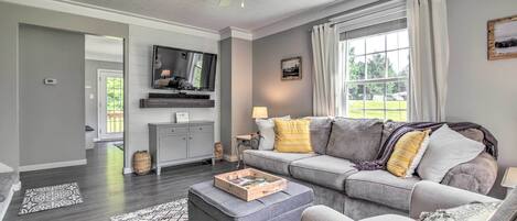 Un salon avec un canapé gris, un pouf et un fauteuil, un ventilateur de plafond, un téléviseur à écran plat et une fenêtre avec des rideaux.