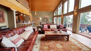Ein geräumiges Wohnzimmer mit einem Ledersofa, einem Couchtisch aus Holz und einem gemusterten Teppich. Eine Treppe führt auf eine obere Ebene, und große Fenster bieten Ausblick ins Freie.