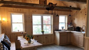 A wooden cabin interior with a kitchen area, large windows, and a bench with pillows. - Lovelyt one bedroom nestled in the white mountains and the lakes region of NH (ORFORD)