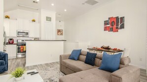 Una sala moderna con un sofá color beige, una mesa de centro blanca y un sillón azul. Se ve una cocina con gabinetes blancos y electrodomésticos de acero inoxidable al fondo.