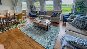 Un salon moderne avec un canapé, une table basse et des fauteuils. De grandes fenêtres sont ornées de rideaux, une plante est présente, et on peut admirer la verdure à l’extérieur.