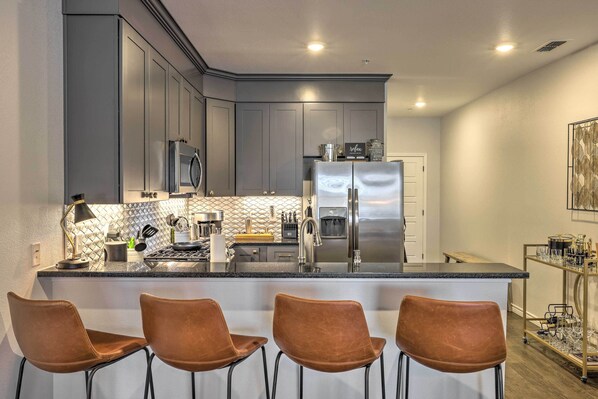 A modern kitchen with grey cabinets, stainless steel appliances, a marble countertop, and four brown leather bar stools.