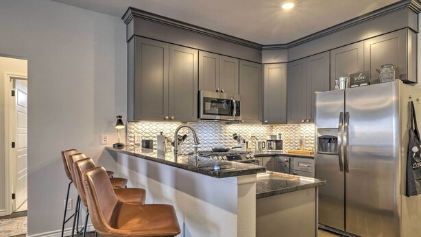 A modern kitchen with grey cabinets, stainless steel appliances, a central island with bar stools, and a tiled backsplash. - Single-Story Condo < 1 Mile From Downtown! (Fredericksburg)