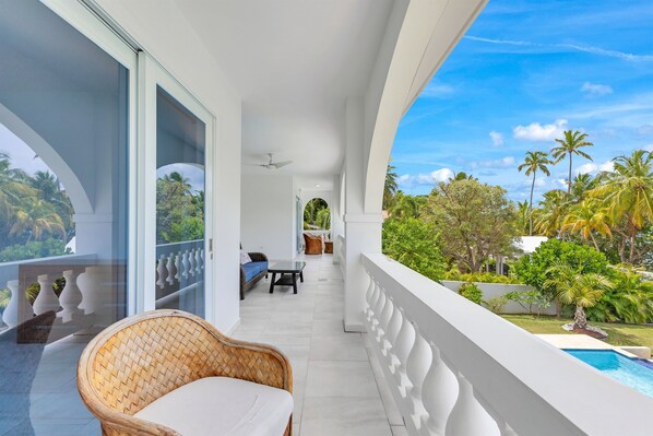 A balcony with a wicker chair, a sofa, and a table. There is a pool visible in the background.