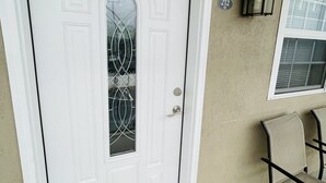 A white door with a glass panel, a doormat, a wall-mounted light fixture, and a chair.