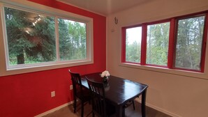 A dining area with a table and chairs, large windows with red frames, and a view of trees outside.