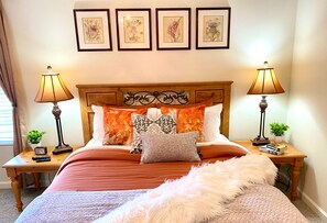 A neatly arranged bedroom with a wooden headboard, multiple pillows, two bedside tables with lamps, and framed artwork on the wall.
