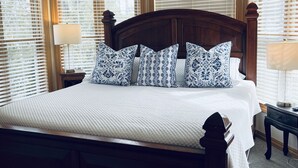 Un grand lit avec une tête de lit en bois, une literie blanche et des coussins à motifs. Il y a deux tables de chevet avec des lampes, et la pièce est dotée de grandes fenêtres équipées de stores.