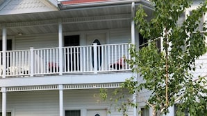 Un bâtiment à deux étages, à la façade blanche, doté d’un balcon à l’étage supérieur et d’un petit jardin devant, agrémenté de diverses plantes et fleurs.