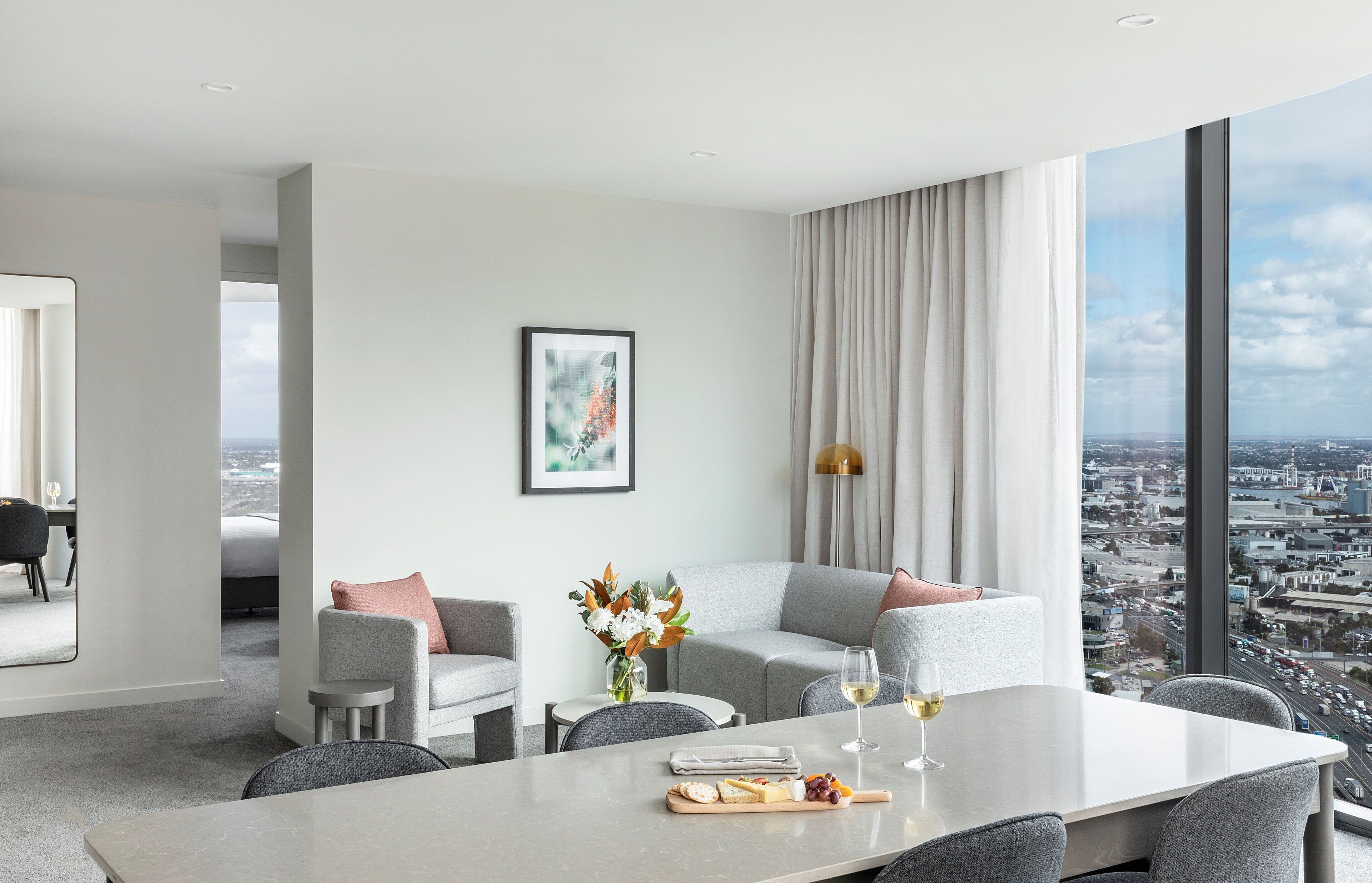 A modern hotel room with a large dining table, grey sofas, a city view, and a framed artwork on the wall.