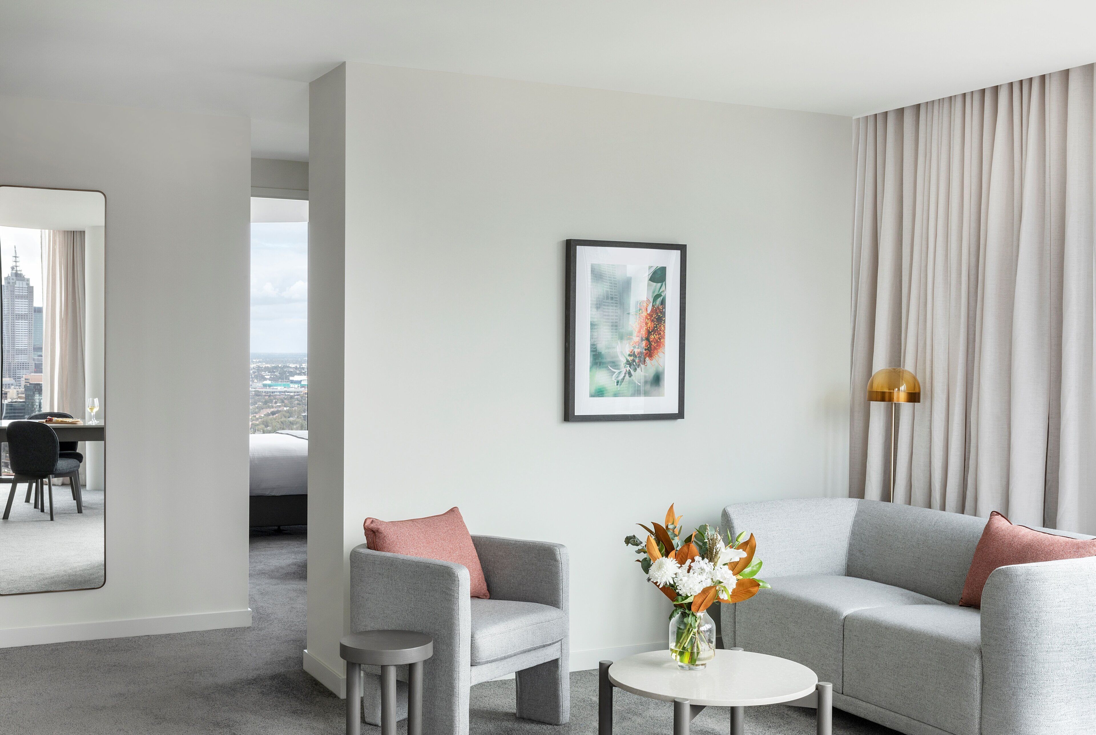A modern hotel room with a grey sofa, a small round table with a vase of flowers, a framed picture on the wall, and a view of a cityscape through a window.