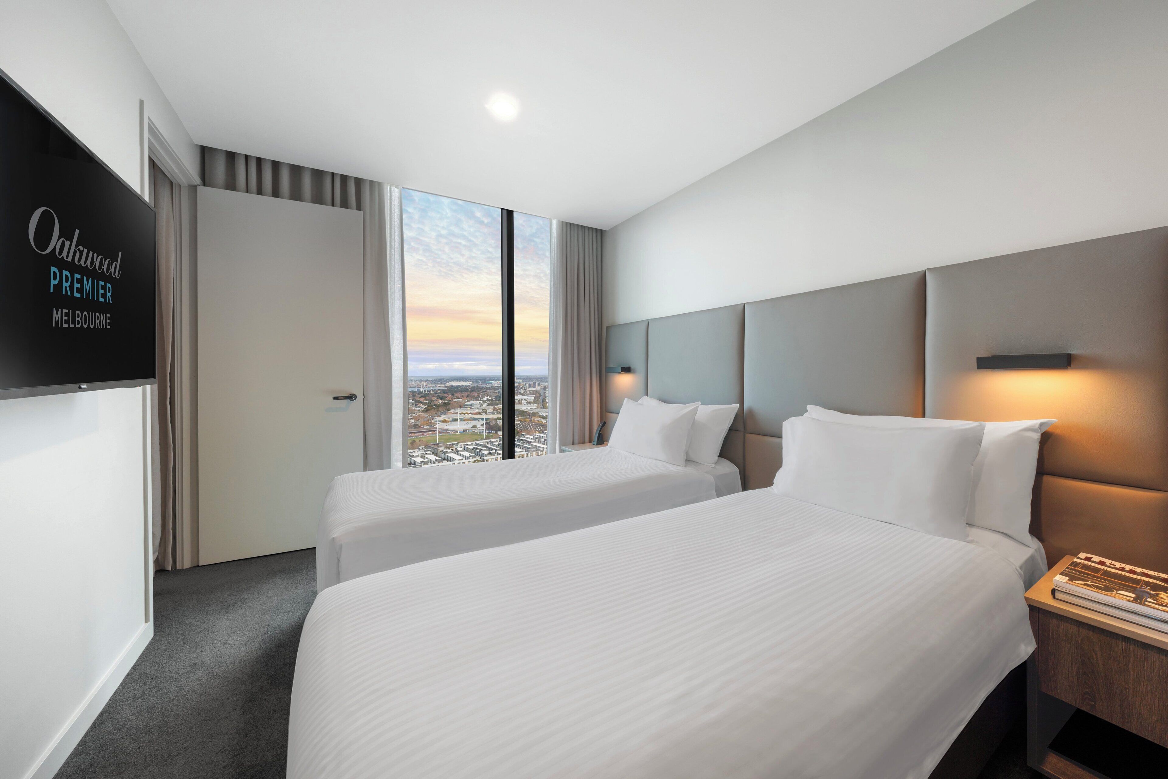 A hotel room with two beds, a TV on the wall, and a view of the city.
