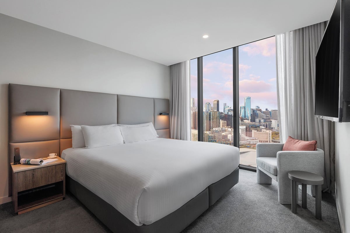A modern hotel room with a large bed, a city view, a flat-screen TV, and a seating area.